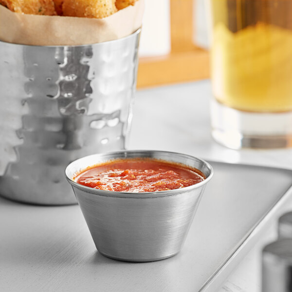 A Tablecraft stainless steel sauce cup filled with red sauce on a table next to fried chicken sticks.