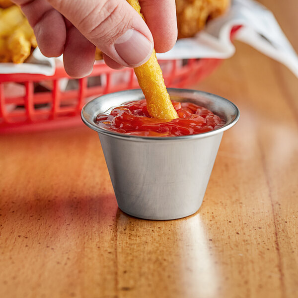 A person dipping a Tablecraft stainless steel sauce cup with ketchup on a french fry.