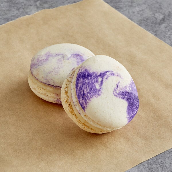 Two Macaron Centrale blackberry cheesecake macarons with purple streaks on them sitting on a piece of paper.