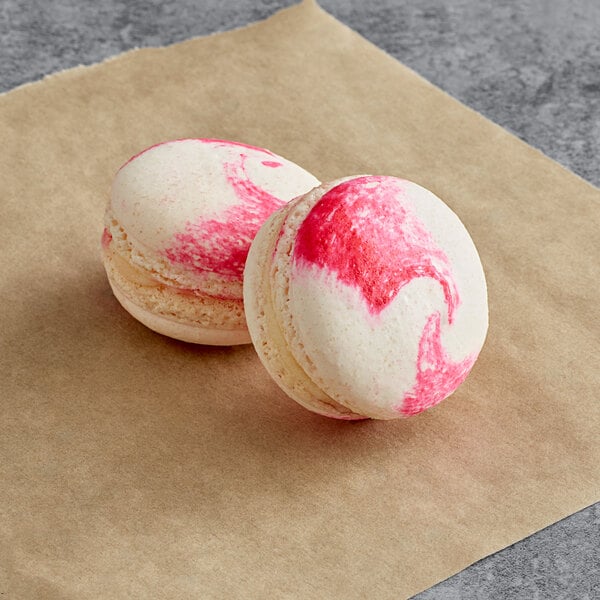 Two Raspberry Cheesecake Macarons with pink and white swirls on brown paper.