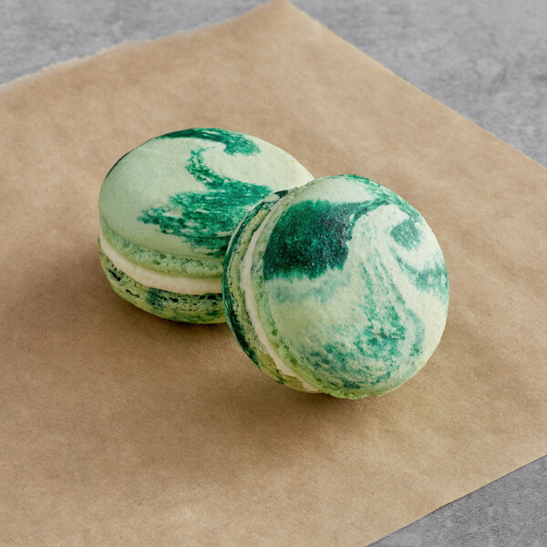Two Macaron Centrale Pina Colada macarons with green and white shells on brown paper.