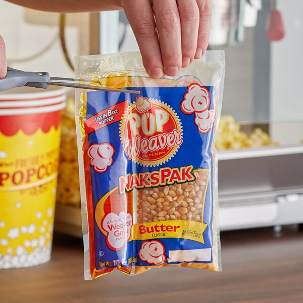 A person holding a Pop Weaver All-In-One Naks Pak Butter Burst popcorn bag in front of a popcorn machine.