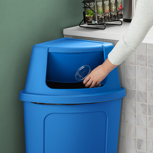 A woman putting a plastic cup into a Lavex blue corner round trash can.