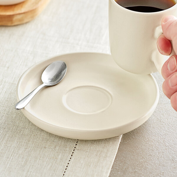 A hand holding an Acopa fog white porcelain cup of coffee on a saucer with a spoon.