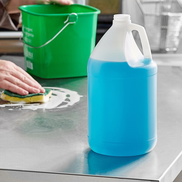 A person cleaning a home kitchen counter with a 1 gallon translucent jug of blue liquid.