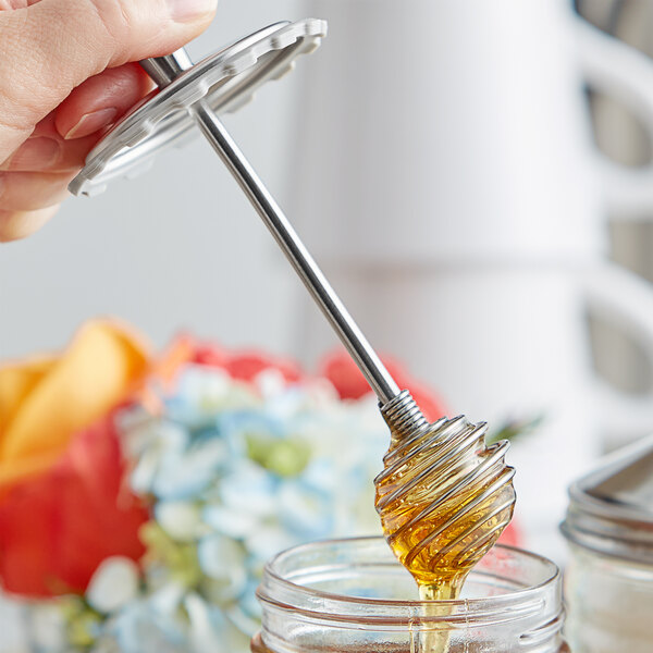A hand holding a Fox Run stainless steel honey dipper over a jar of honey.