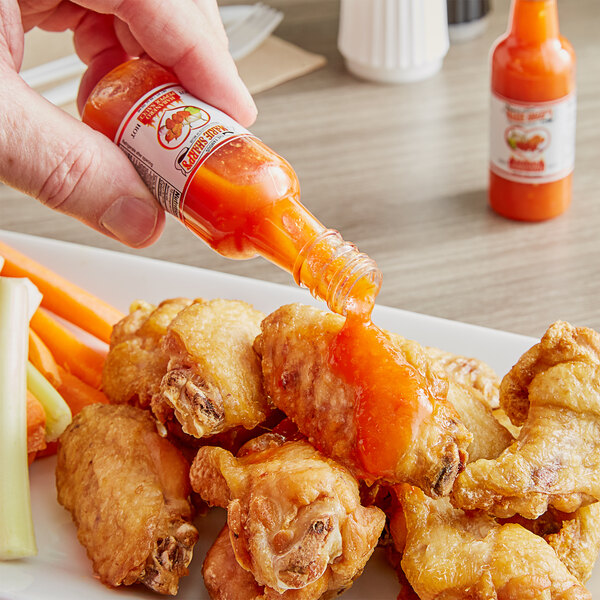 A hand pouring Marie Sharp's Hot Habanero Hot Sauce onto a plate of chicken wings.