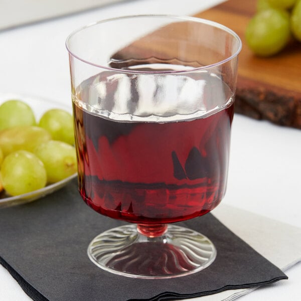 A Fineline clear plastic wine cup filled with red liquid next to grapes.