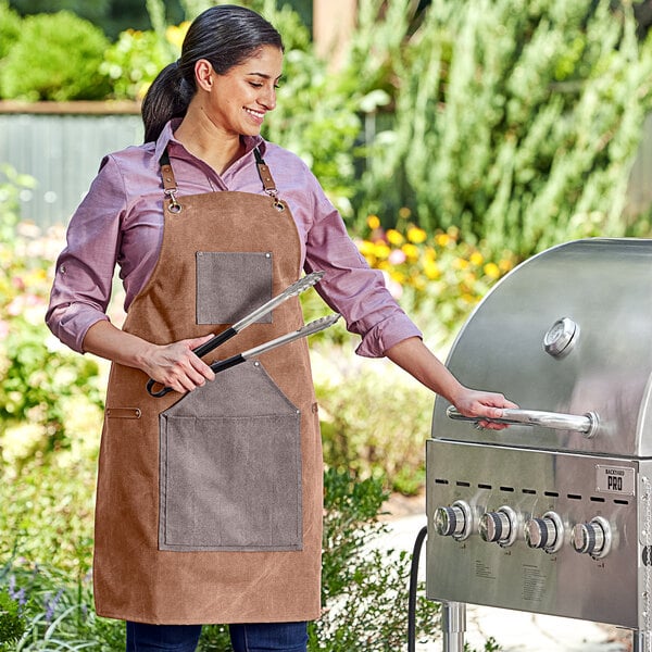 A woman wearing a Backyard Pro heavy-duty canvas grilling apron and holding tongs to a grill.