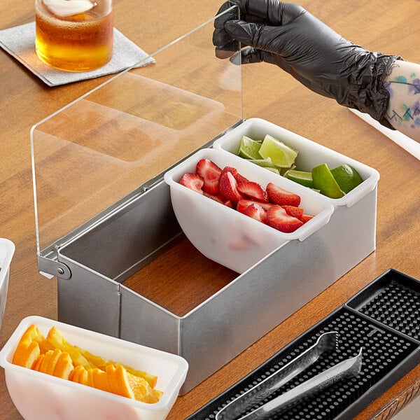 A person using a Choice stainless steel condiment bar to put fruit in a white container.