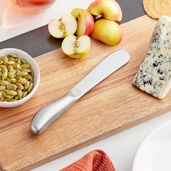 An Acopa stainless steel cheese spreader on a cutting board with cheese and fruit.