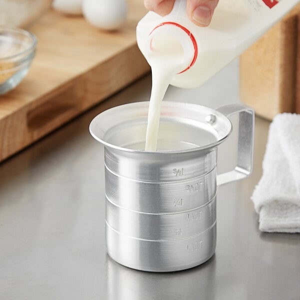 A hand pouring milk into a Choice aluminum measuring cup.