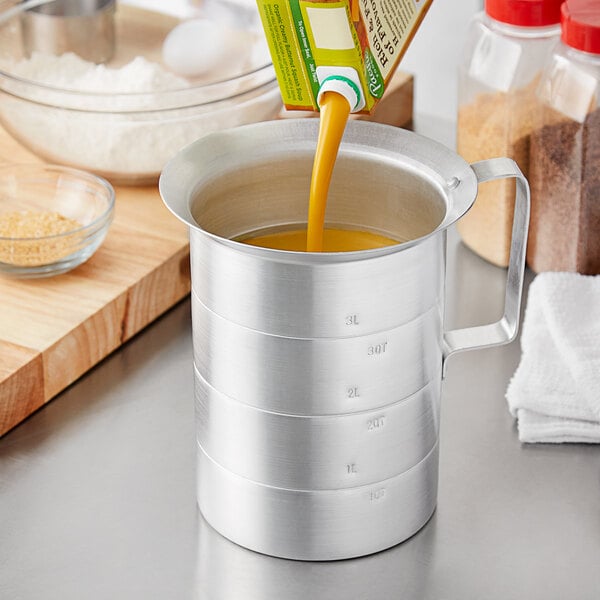 A person pouring orange juice from a carton into a metal measuring cup with a handle and pour lip.