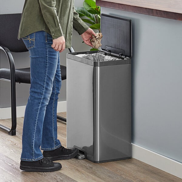 A person using a Rubbermaid charcoal stainless steel step-on trash can.