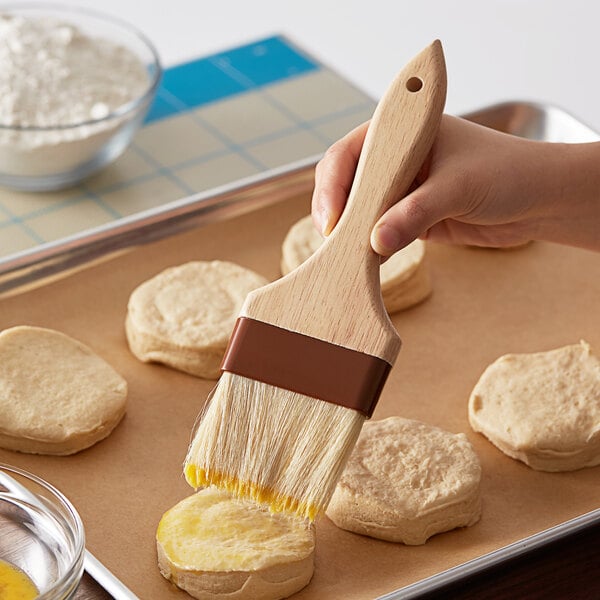 A person using a 3" wide boar bristle pastry brush with a wood handle to brush biscuits.