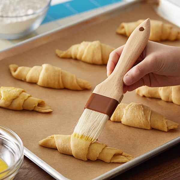 A person using a 1 3/8" Boar Bristle Pastry Brush with a wooden handle to brush croissants on a tray.