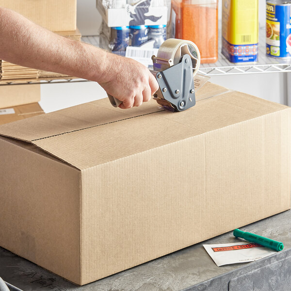 A hand using a tape gun to seal a Lavex Kraft cardboard shipping box.