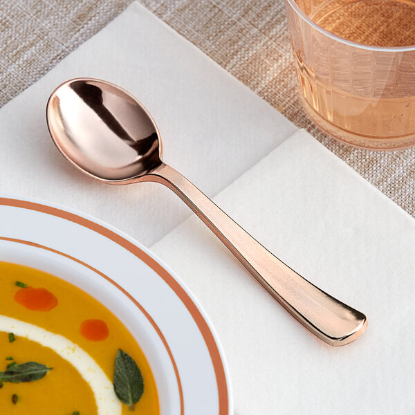 A Visions rose gold plastic soup spoon on a plate next to a glass of liquid.
