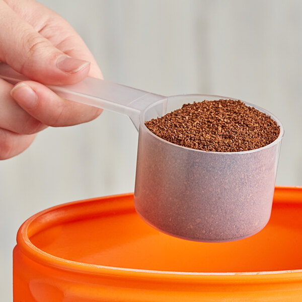 A hand using a 90 cc polypropylene scoop to pour brown granules into a container.