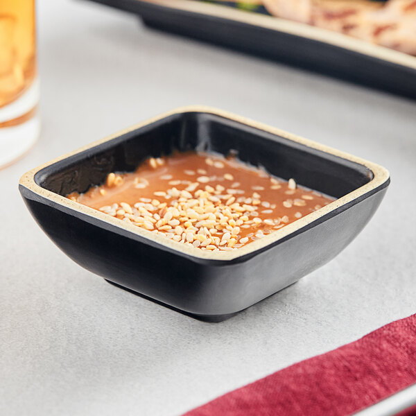 An Acopa matte black square melamine ramekin with ivory rim filled with brown sauce on a table in an Asian restaurant.