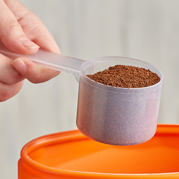 A hand using a 94 cc polypropylene scoop with a long handle to pour brown powder into a container.