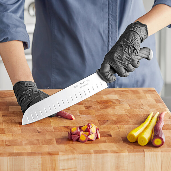 A person using a Schraf Santoku knife to cut a yellow and purple carrot on a cutting board.