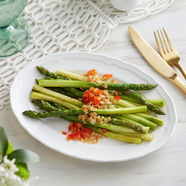 A pearl white Acopa Condesa porcelain platter with asparagus and tomatoes on a table with a fork and knife.