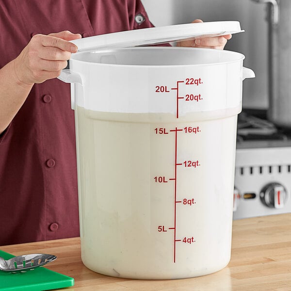 A woman using a measuring cup to pour liquid into a large white Choice food storage container with a lid.
