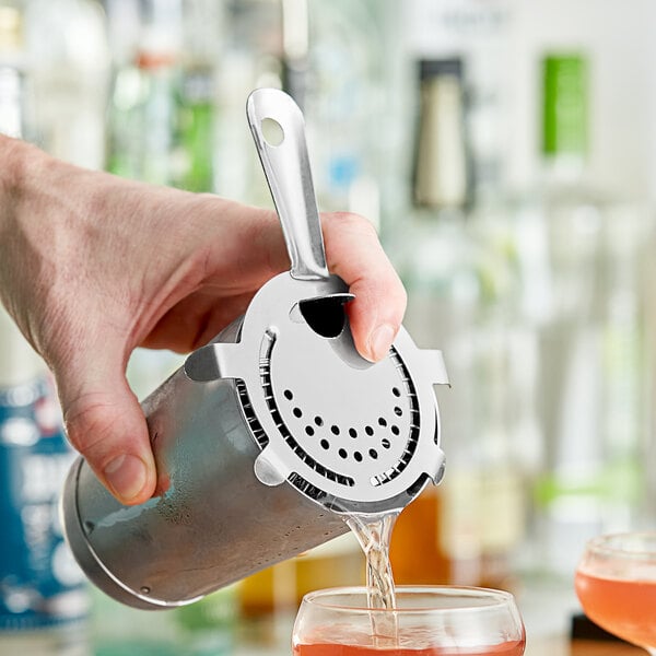 A hand using a Choice stainless steel Hawthorne strainer to pour a cocktail from a silver container.