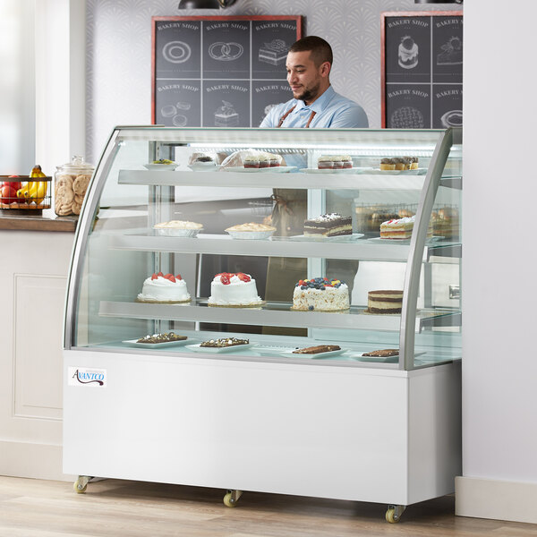 An Avantco refrigerated bakery display case with cakes on shelves and a curved glass front, with a man in a blue shirt behind it.