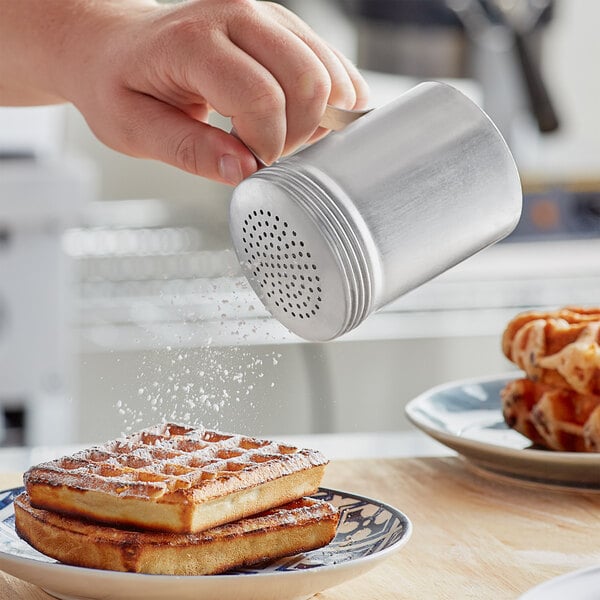 A person holding a Choice aluminum shaker filled with powdered sugar and pouring it onto a waffle.