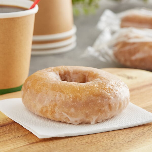 An individually wrapped glazed donut on a napkin next to a cup of coffee.