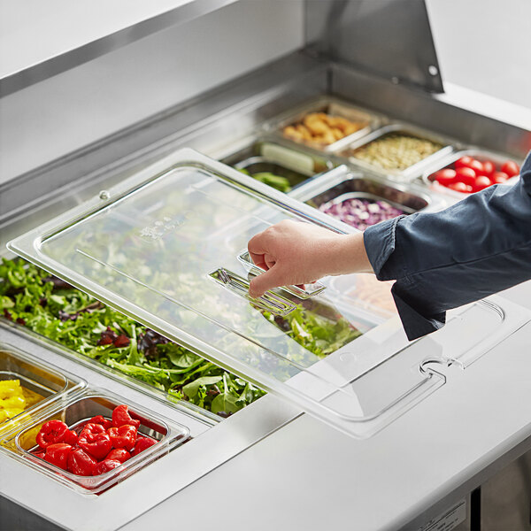 A person's hand reaching into a Cambro clear plastic lid with a spoon notch over a salad bar.