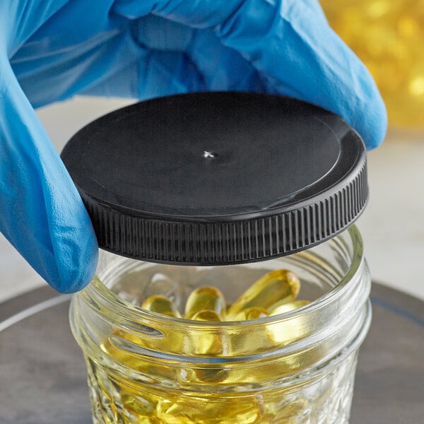 A hand in a blue glove holding a jar with a black 70/400 plastic cap containing yellow pills.