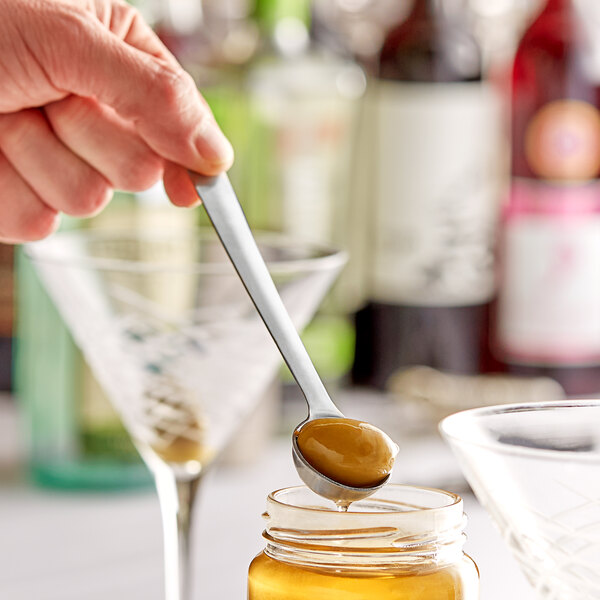 A hand using a Franmara stainless steel olive spoon to pour honey into a martini glass.