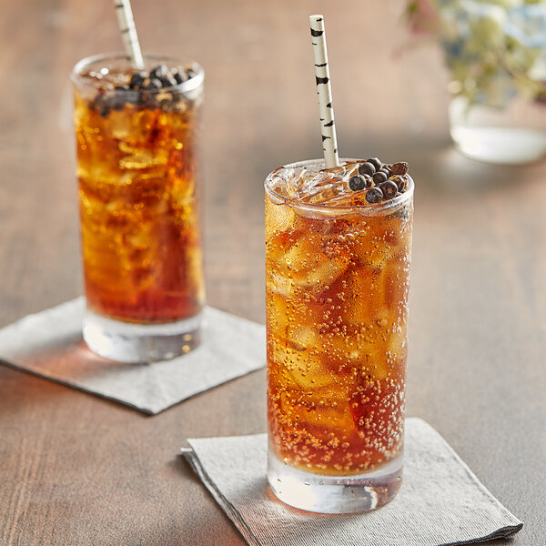 A glass of Tractor Beverage Co. Organic Root Beer soda with ice and a straw next to another glass of the same soda with ice and a straw.