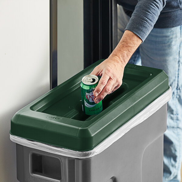 A hand opening a green and white Toter Slimline trash can lid to put a can in it.