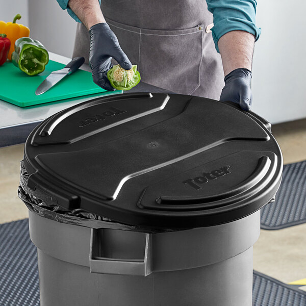 A man in a black apron and gloves putting a black lid on a Toter 32 gallon round trash can.