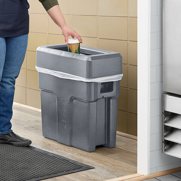 A woman pouring water from a bottle into a dark gray Toter Slimline trash can.