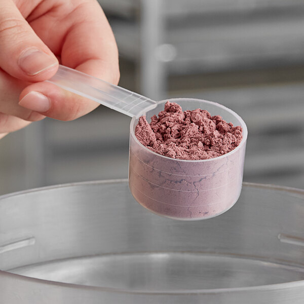 A hand using a long plastic scoop to measure pink powder into a small container.