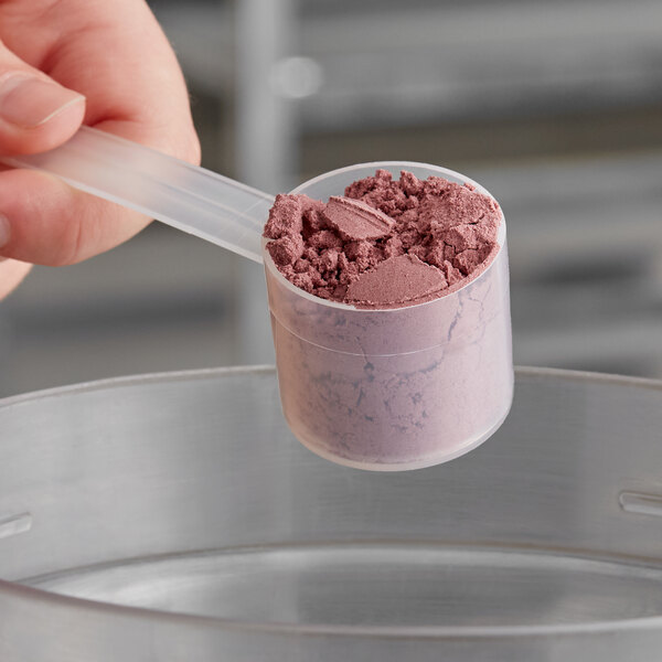 A hand using a long-handled polypropylene scoop to measure pink powder into a clear plastic container.