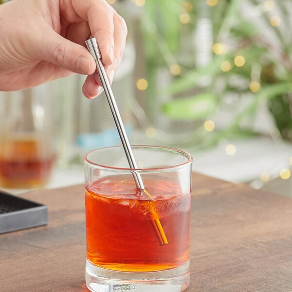 A hand holding a Franmara stainless steel bar stirrer in a glass of brown liquid.
