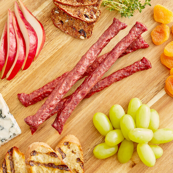 A wooden cutting board with sweet and smokie Shaffer Venison Farms snack sticks, dried apricots, dried fruit, and sliced red apples.