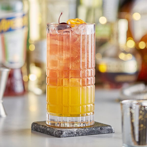 A close-up of an Acopa Madras beverage glass filled with a yellow cocktail and garnished with a cherry and orange slice on a stone coaster.
