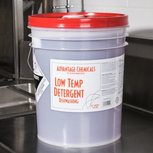 A bucket of Advantage Chemicals low temperature dish washing machine detergent on a counter.