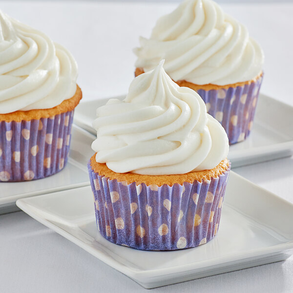 Three Enjay purple fluted baking cups with white polka dots filled with cupcakes with white frosting on a white plate.
