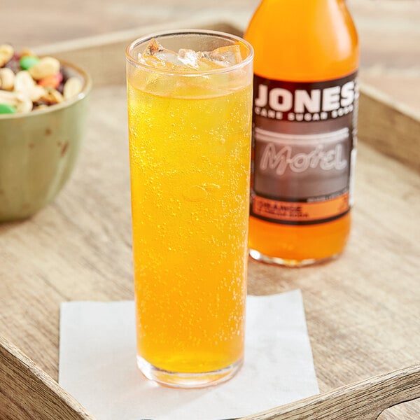 A close up of a glass of Jones orange soda next to a bowl of nuts and a bottle.
