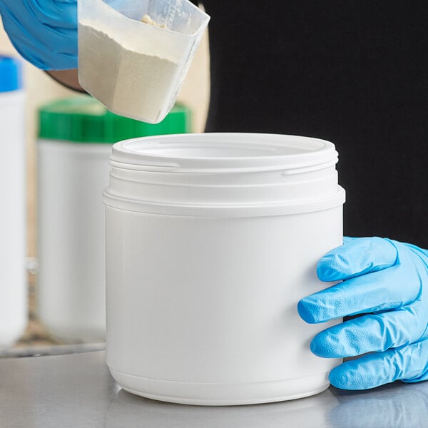 A person in blue gloves pouring white powder into a 44 oz. white plastic container.