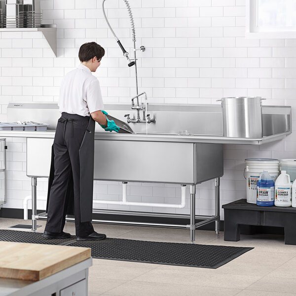 A person in a professional kitchen cleaning a Regency stainless steel three compartment sink.