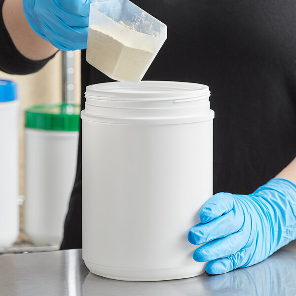 A person in blue gloves pouring white powder into a white plastic container.